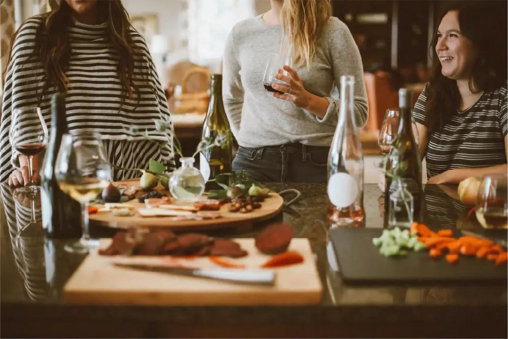 Several women standing around a table full of snacks, talking and laughing