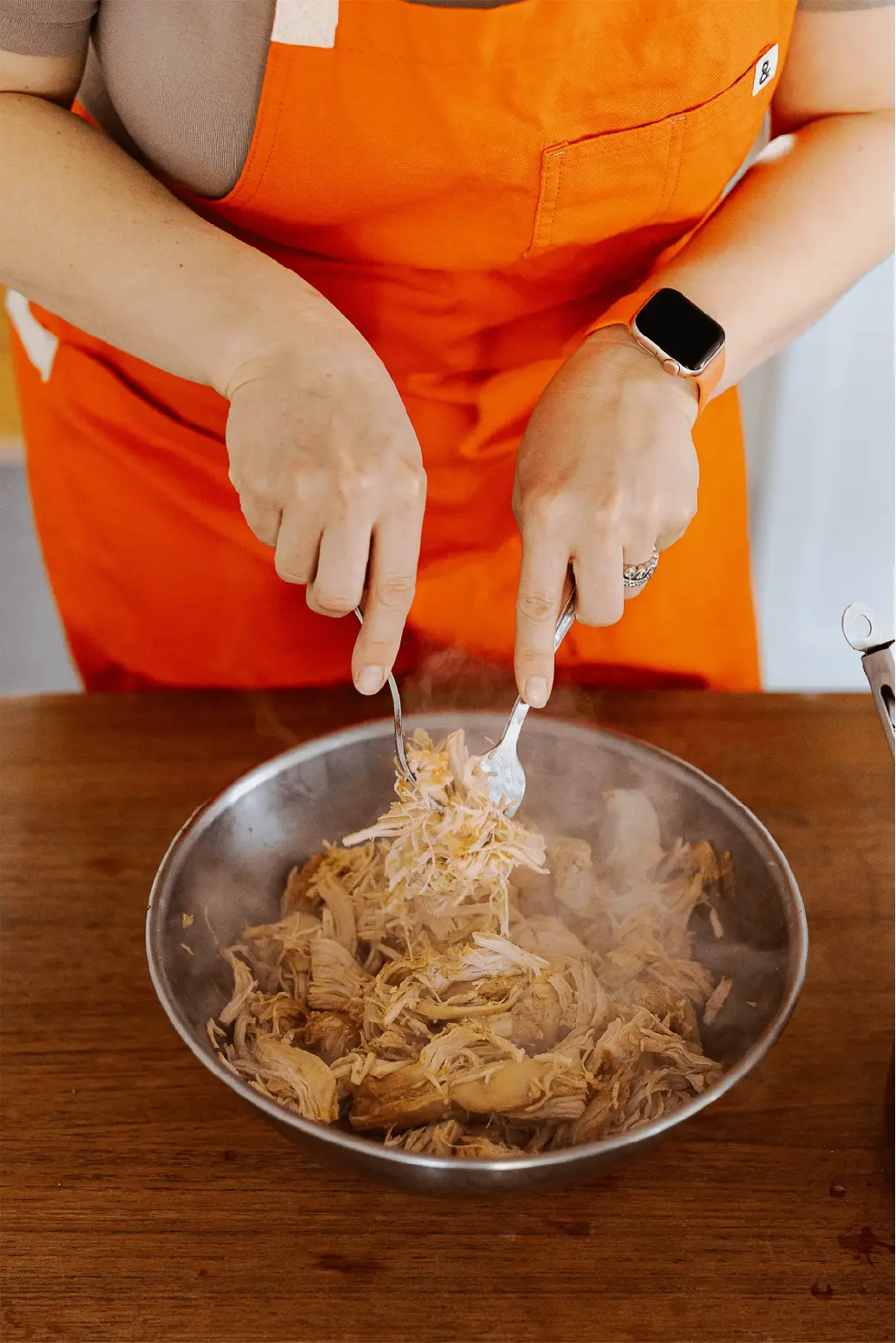 A person in an orange apron pulling apart slow cooked chicken with two forks in a metal bowl