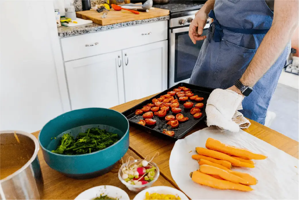 Person wearing a blue apron preparing carrots and tomatoes in a bright kitchen
