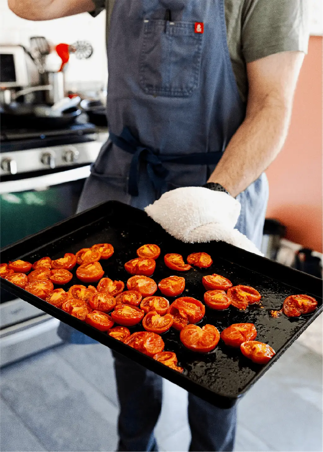 Chef wearing a blue apron holding a black sheet tray filled with cut tomatoes and a white towel